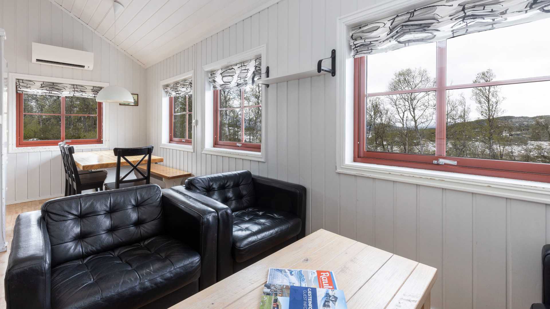 Koselig stue i hytte med svarte sofaer, spisebord og store vinduer med utsikt mot skog og fjell.