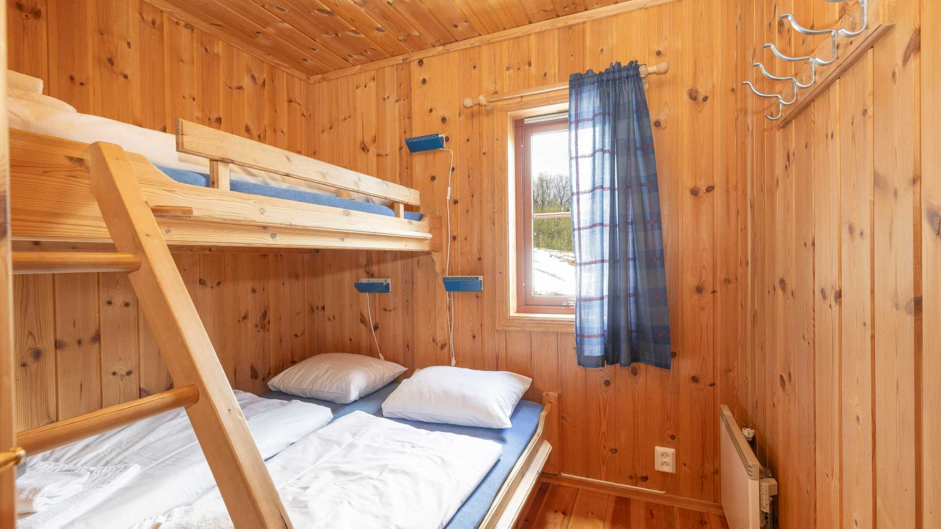 Koselig soverom i trehytte med køyeseng, blå gardiner og naturlig lys fra vinduet.