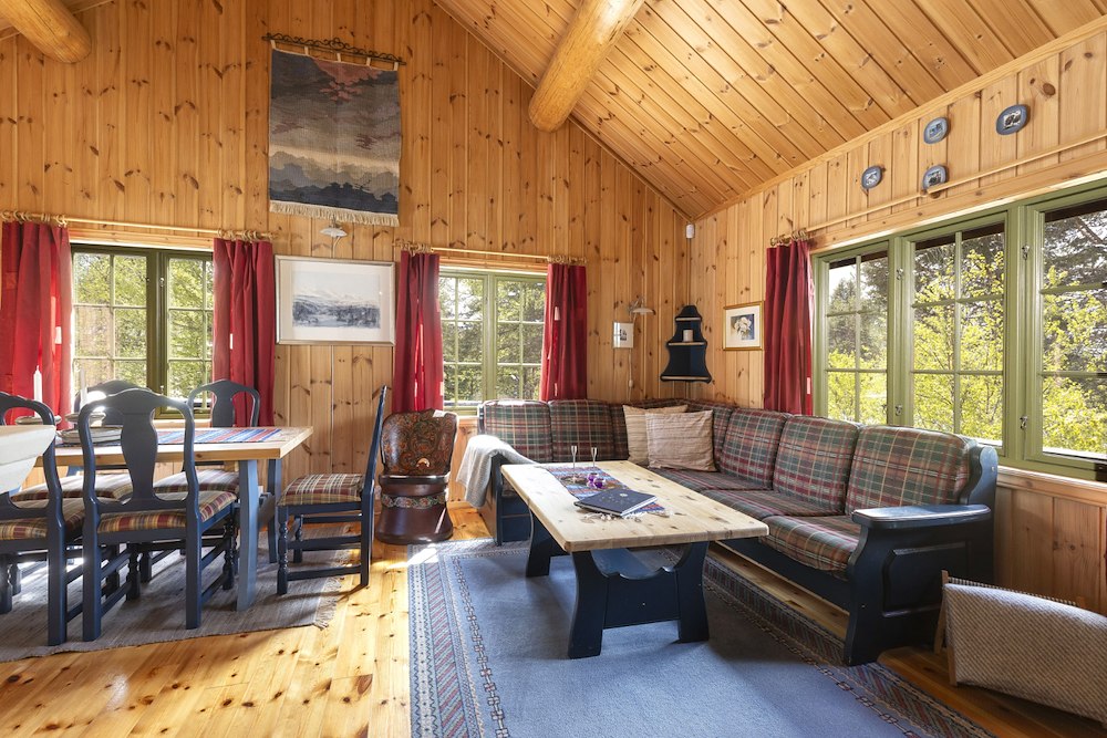 Koselig norsk hytteinteriør med trebjelker, stor sofa, spisebord og vakker utsikt gjennom vinduene.
