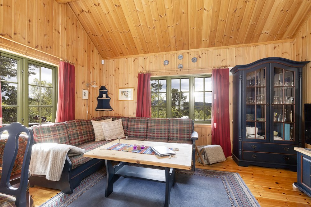 Koselig norsk hyttekrok i treverk med rutete sofa, bord og vinduer som viser skog utenfor.
