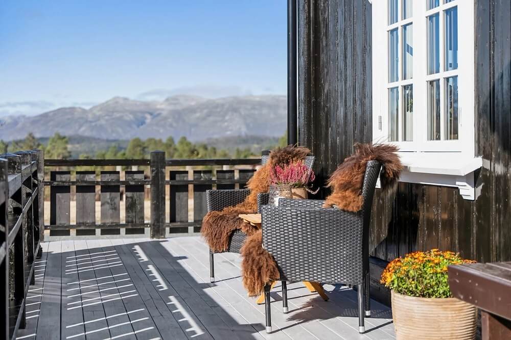 Utsikt over fjell fra terrasse med to stoler og bord, dekorert med pelser og blomster, solfylt dag.