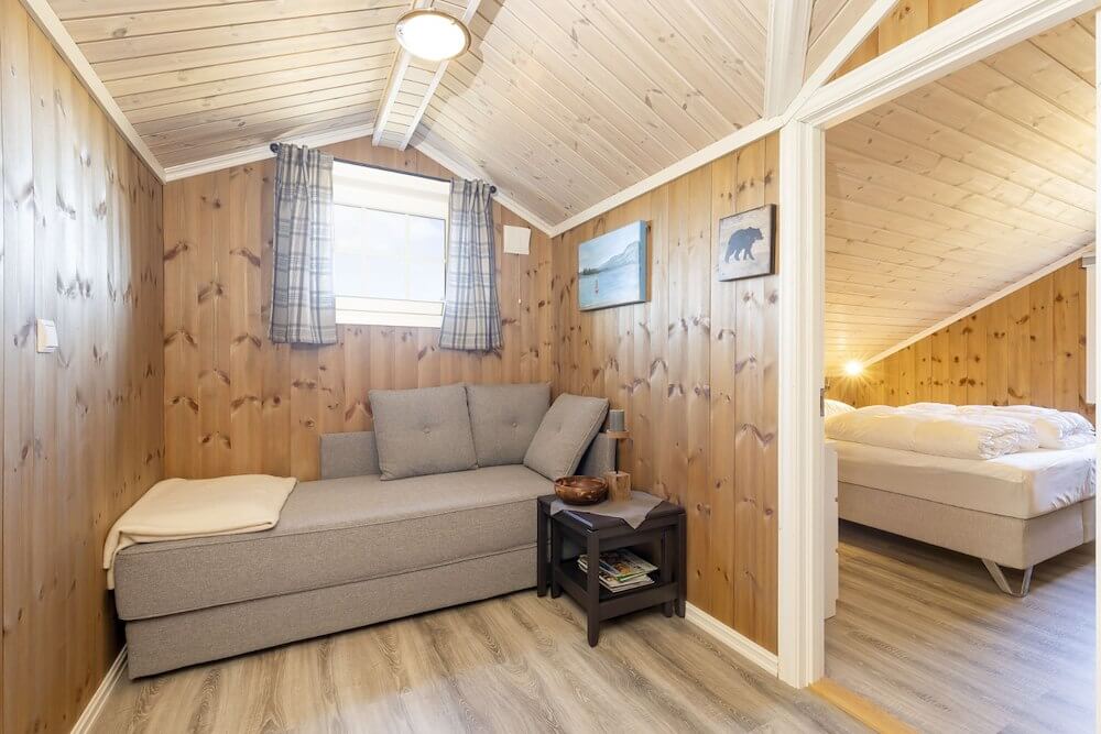 Koselig stue i trehytte med komfortabel sofa og soverom i bakgrunnen, norsk fjellhytteinteriør.