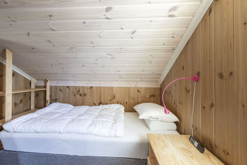 Loftseng med trepaneler og leselampe i rosa, koselig soverom i norsk hyttestil.