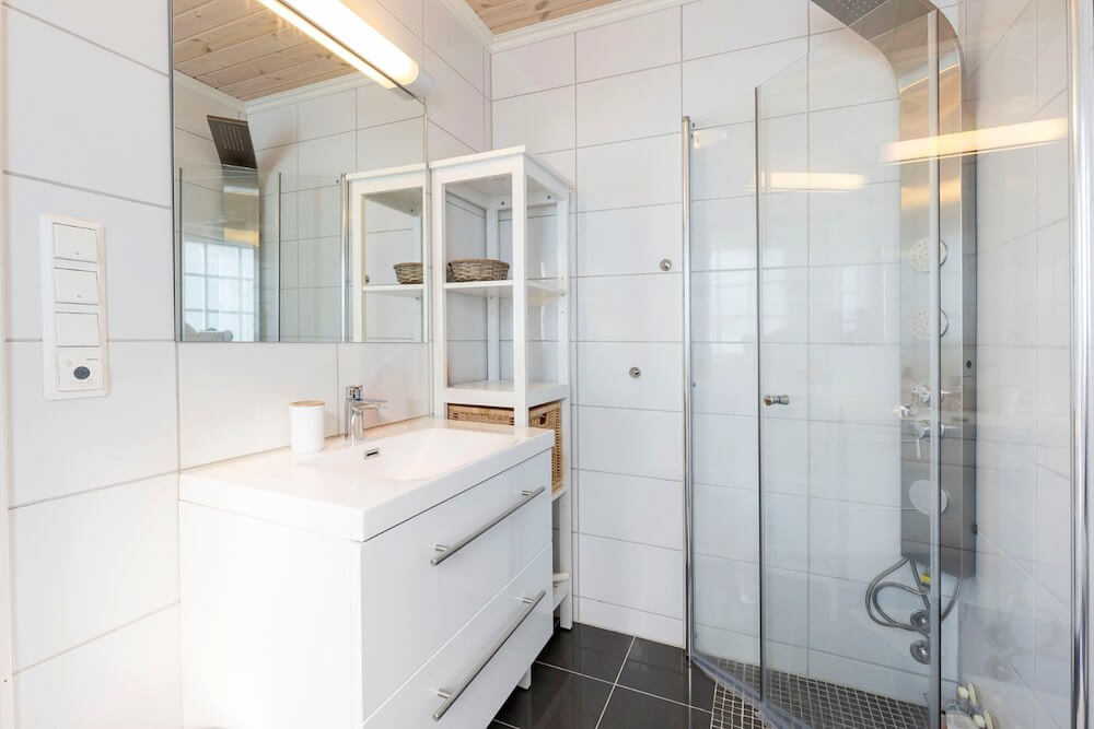 Lyst og moderne bad med dusjkabinett, hvit servant og speilskap på hvite fliser.