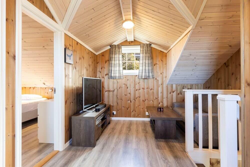 Loftstue med trepanel, TV, sofagruppe og stort vindu. Seng i naborommet. Hyggelig og lys atmosfære.