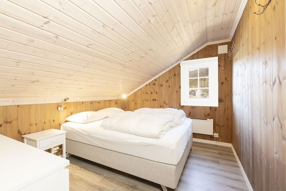Lyst soverom med skråtak, dobbeltseng, trepanelvegger og et vindu som slipper inn naturlig lys.