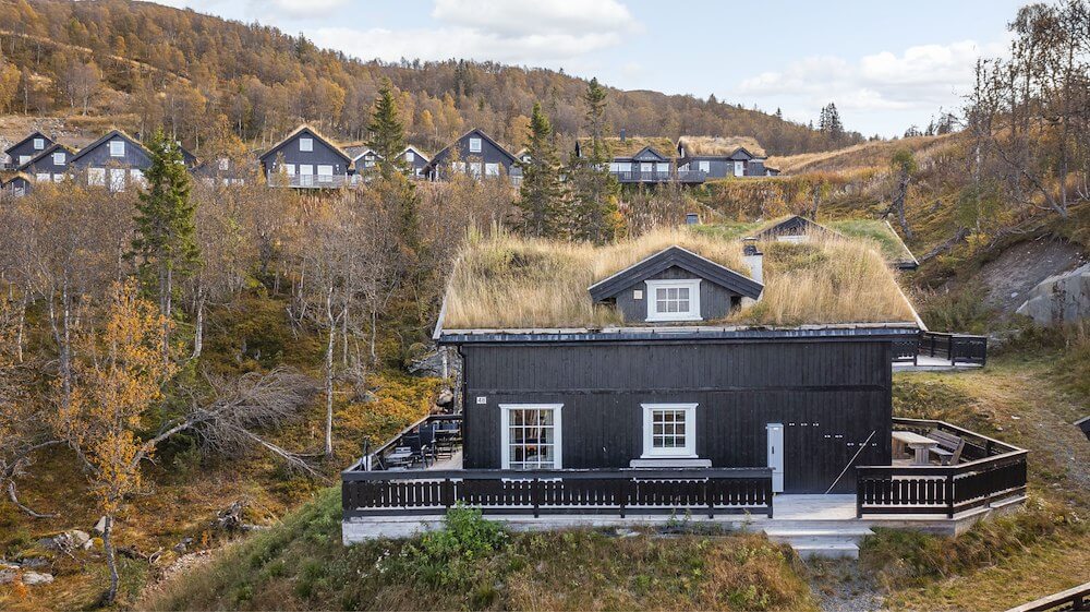 Svart hytte med torvtak i norsk fjellandskap, omgitt av høstfarger og skog, med flere hytter i bakgrunnen.