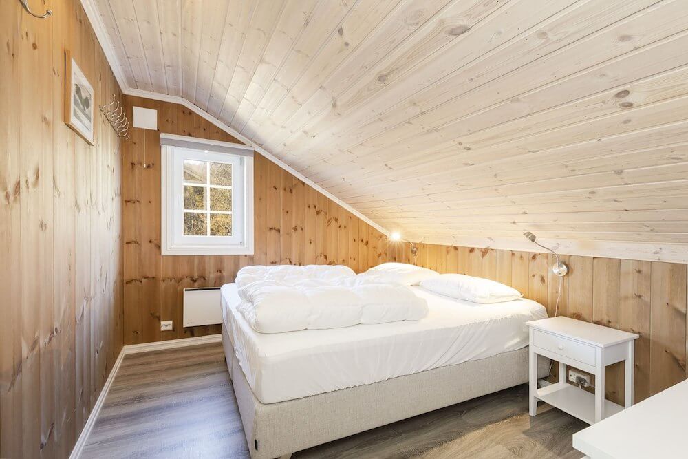 Koselig soverom med trepanel, skråtak, hvit seng og nattbord, vindu med utsikt og innbydende atmosfære.