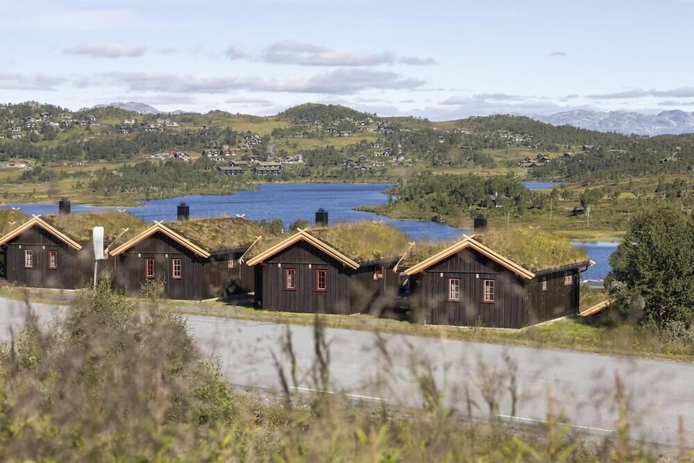 Tre norske hytter med torvtak ved innsjø og grønne åser under blå himmel.