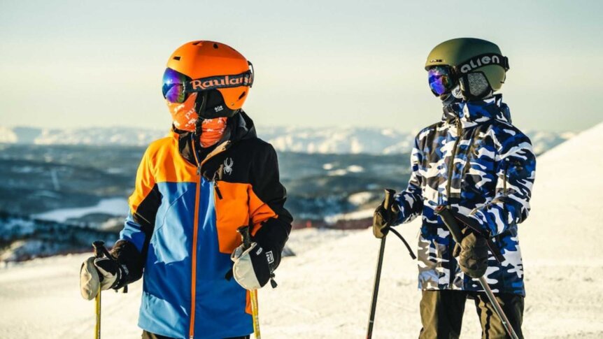 To personer kledd for skisport i fargerike jakker på snødekt fjell med solfylt bakgrunn.