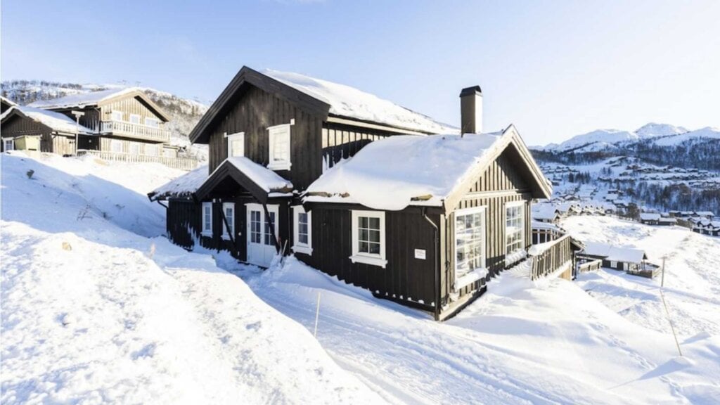 Tradisjonell norsk hytte om vinteren, dekket av snø, med fjellandskap i bakgrunnen.