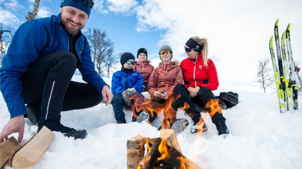 Familie på vintertur nyter bål i snøen med skiutstyr i bakgrunnen, under en klar blå himmel.