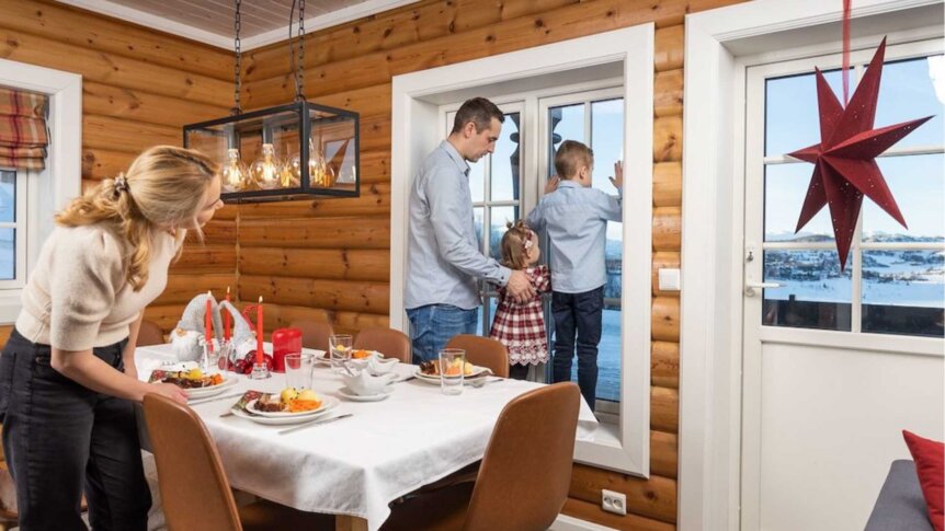 Familie i norsk hytte ser ut vinduet, klar for julemiddag ved pyntet bord med lys og dekor.