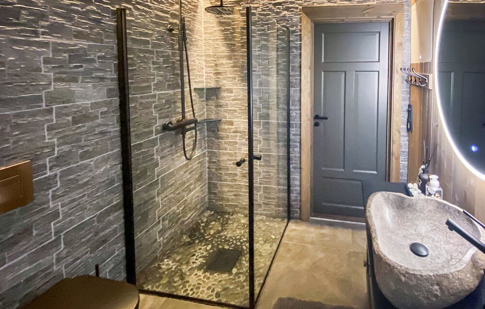 Moderne bad med steinfliser, dusjhjørne i glass og rund speil med LED-lys i et stilrent design.