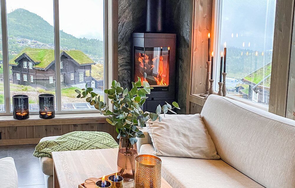 Koselig stue med peis og utsikt over fjellhytter. Store vinduer gir lys til interiøret med planter og lys.