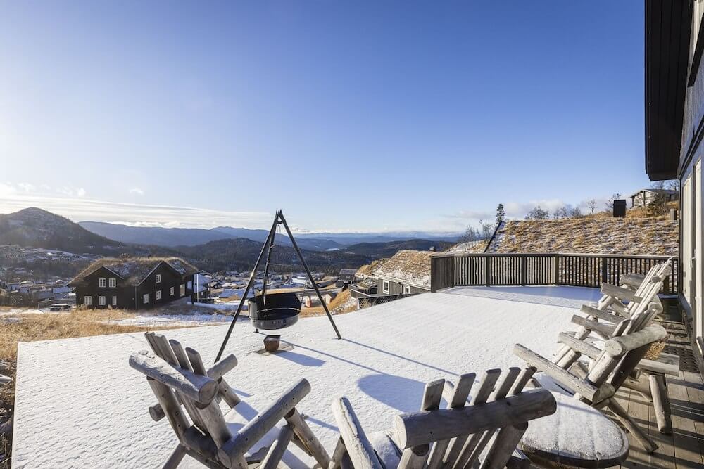 Snødekt terrasse med utsikt over fjellandskap og hytter under en klar blå himmel.