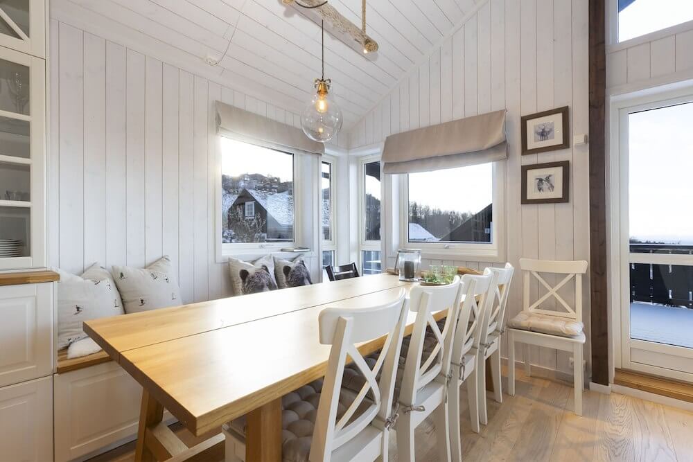 Koselig spisestue i hytte med store vinduer, trepanel og hvite møbler. Utsikt mot vinterlandskap utenfor.