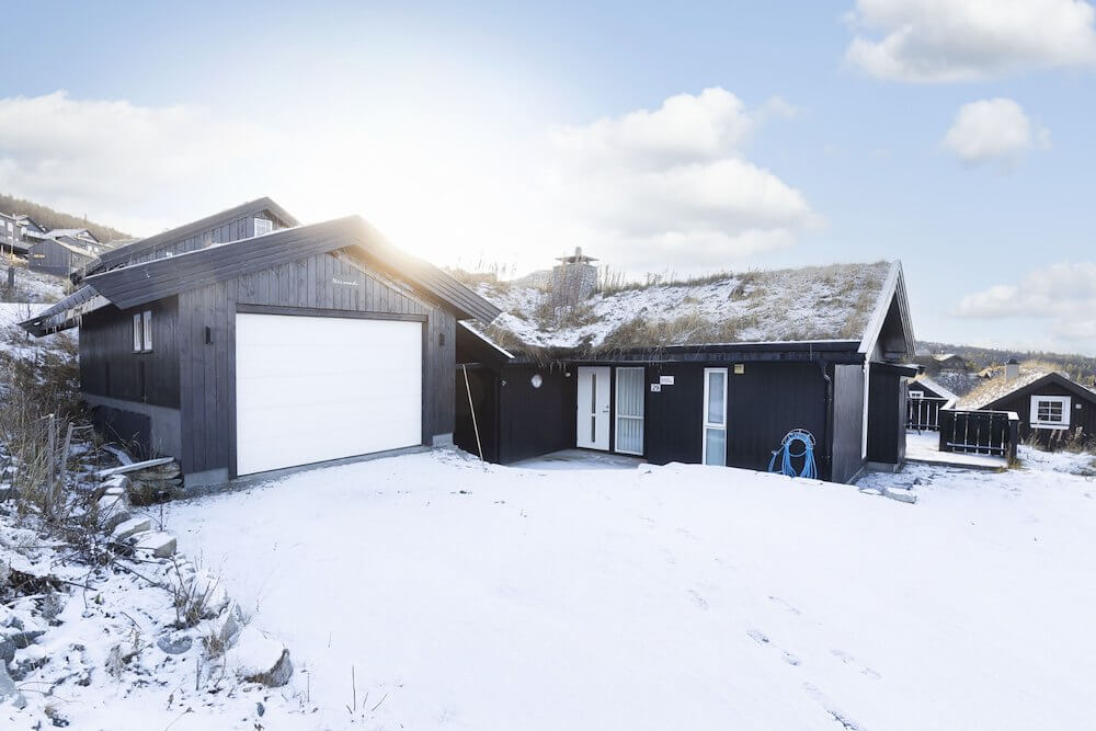 Moderne hytte med torvtak og garasje i snøbelagt landskap, solfylt vinterdag i Norge.