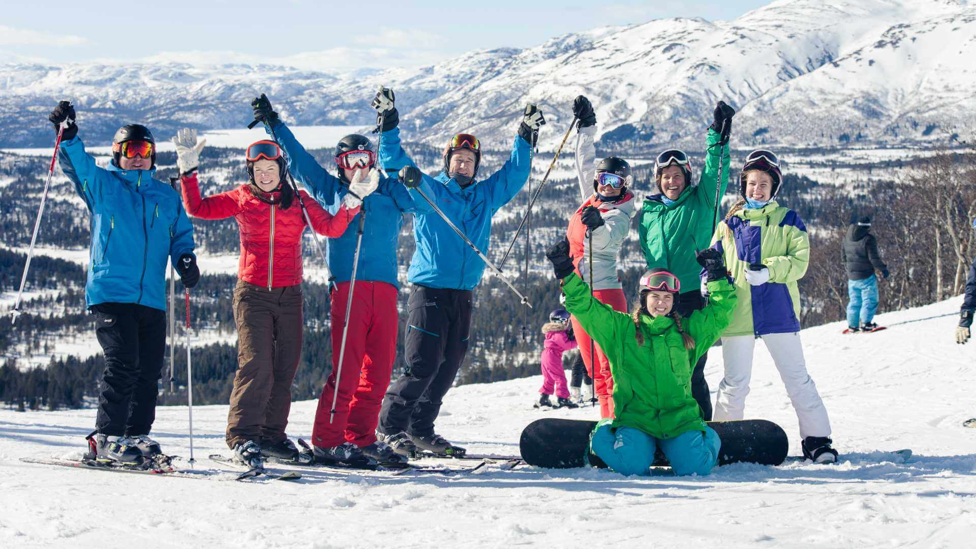 Gruppe på ski og snowboard i snødekte fjell, smilende og glade i solfylt vinterlandskap.