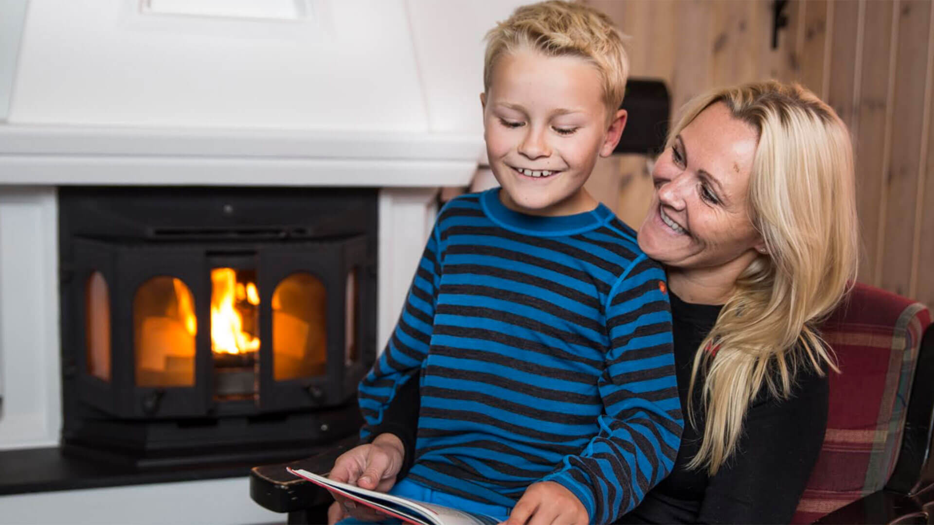 Mor og sønn leser foran en peis, koselig atmosfære med lys og varme.