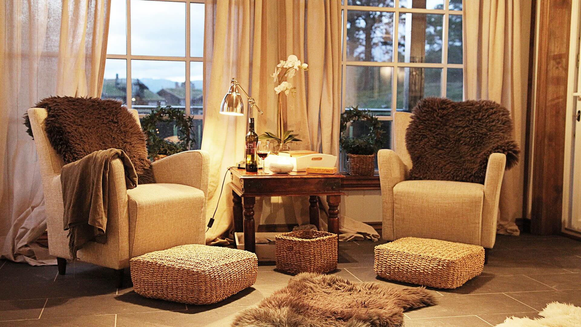 Koselig stue med to lenestoler, saueskinn, trebord og myke koseputer foran store vinduer.