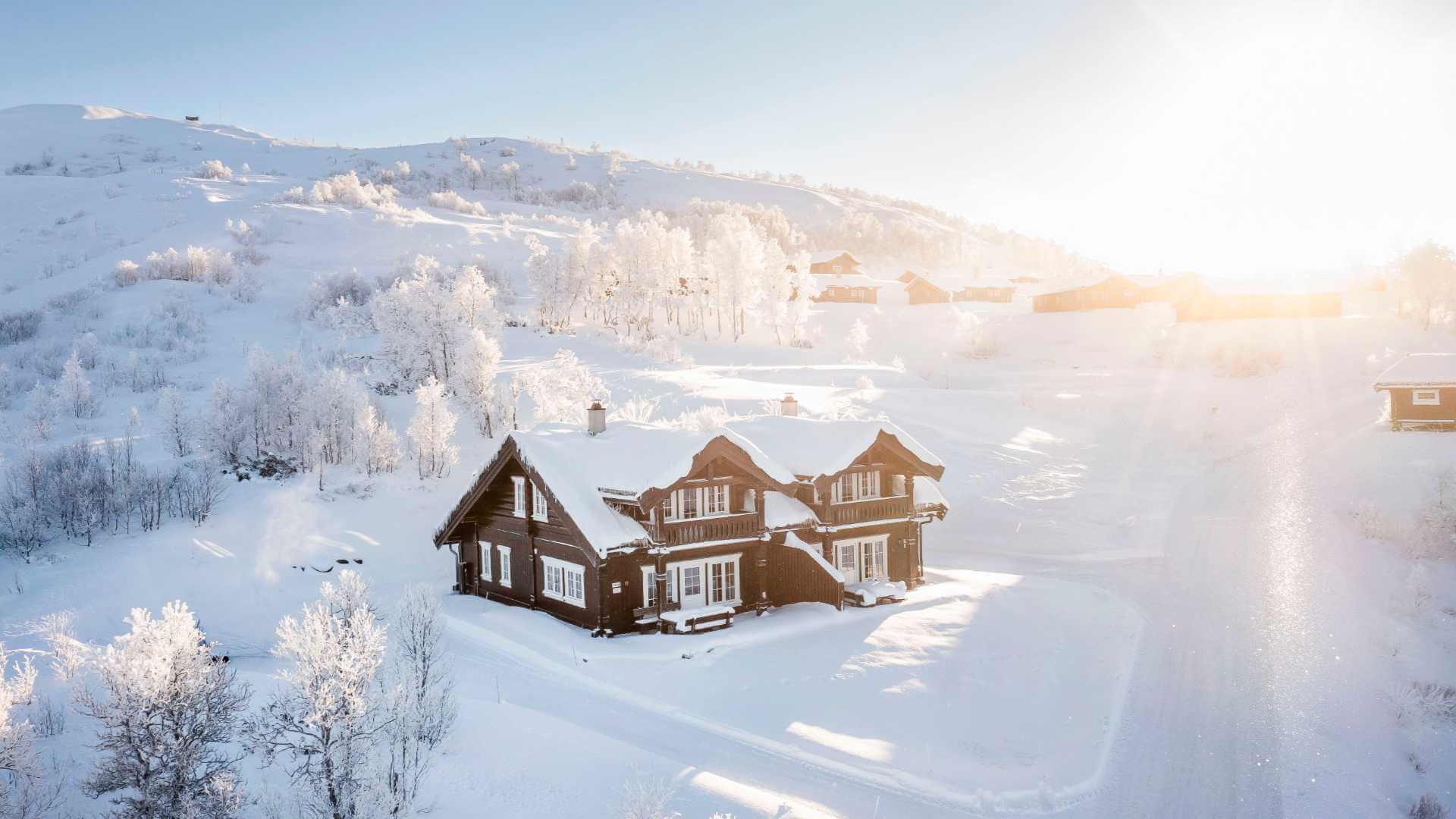 Snødekket hytte i solfylt vinterlandskap med trær og bakke i Norge.