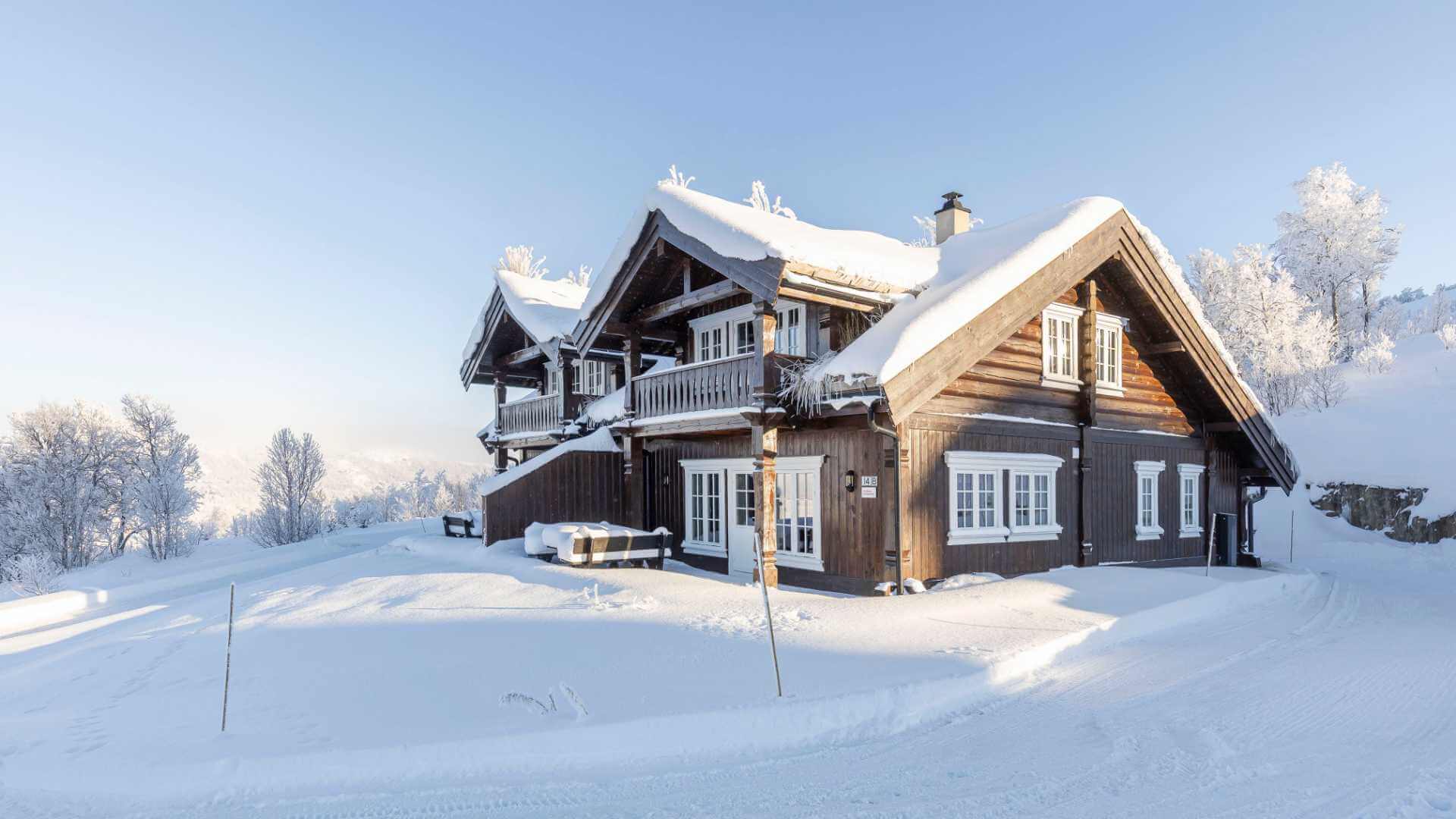 Snødekt norsk hytte i vinterlandskap, omgitt av trær og klar blå himmel.