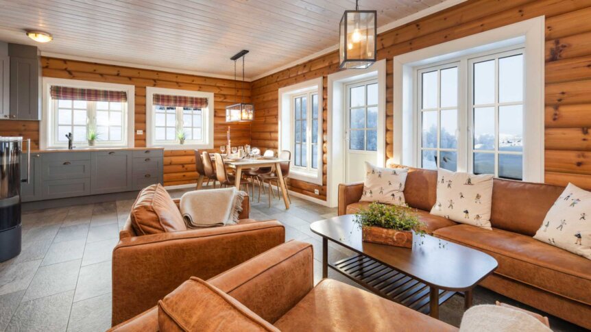 Koselig tømmerstue med brune skinnsofaer, spisestuebord og store vinduer med utsikt.