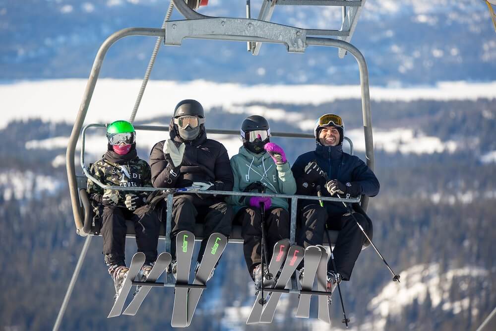 Fire personer på skiheis med skiutstyr, omgitt av vinterlandskap. Klar blå himmel og snødekte trær i bakgrunnen.