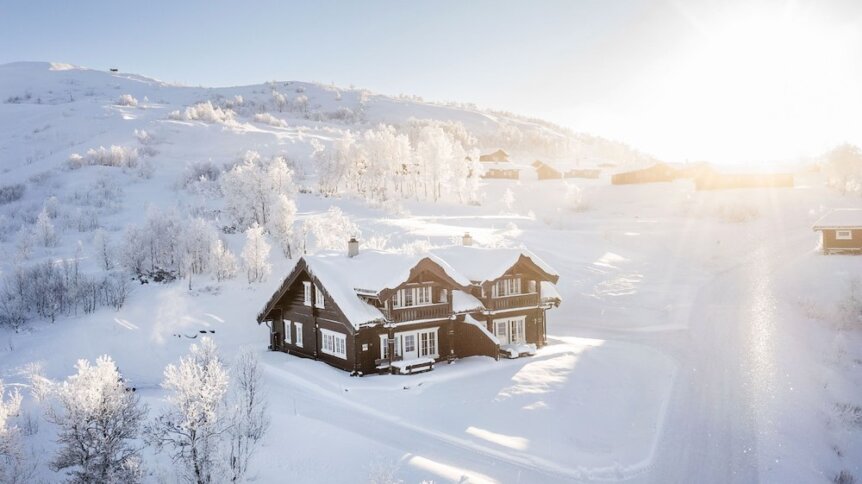 Snødekt norsk fjellandskap med tradisjonell hytte i solskinn, omgitt av vinterkledde trær.