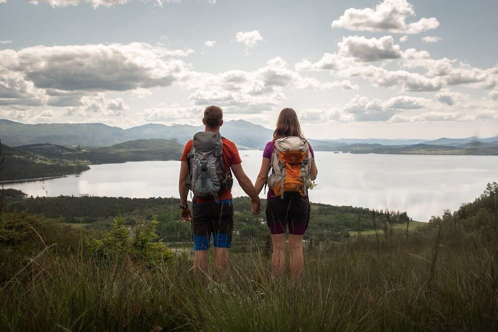 Par på tur holder hender, ser utover vakker natur med innsjø og fjell i bakgrunnen. Ideelt for friluftsliv.