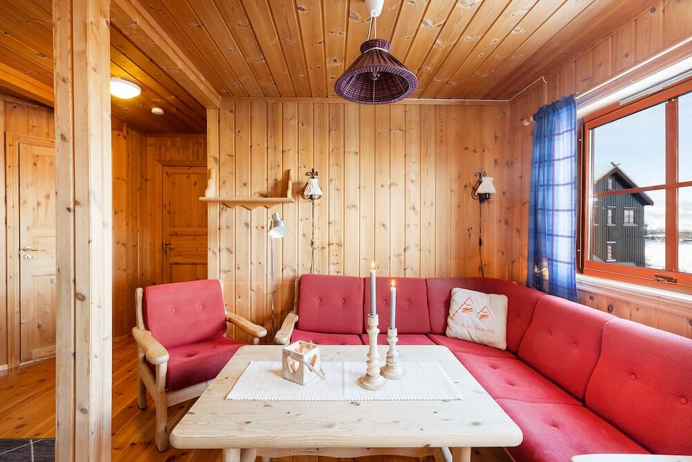 Koselig stue med rød sofa, trepanel, vindu med utsikt og levende lys på bordet, perfekt for avslapning.