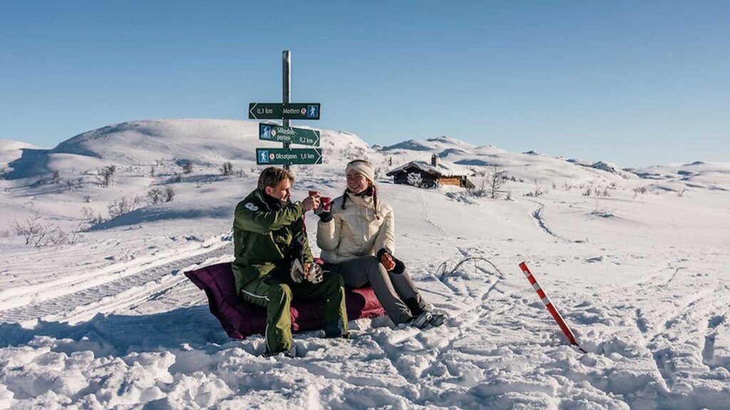 Par med varm drikke sitter på snødekt bakgrunn ved skilt for turgåing i fjellet. Klar himmel og fjelllandskap rundt.