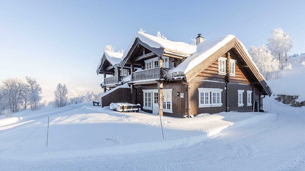 Vakker, snødekt hytte i fjellet om vinteren, omgitt av trær og klar himmel. Ideal for vinterferie og naturopplevelser.