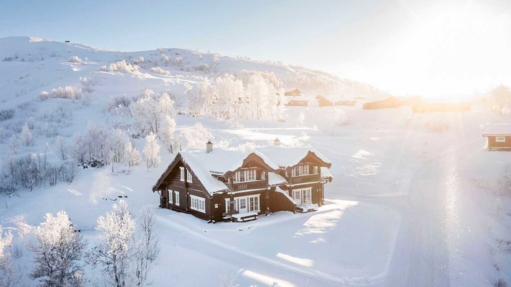 Nydelig hytte i snødekt landskap, omgitt av frostrøde trær og solens stråler som skinner i bakgrunnen.