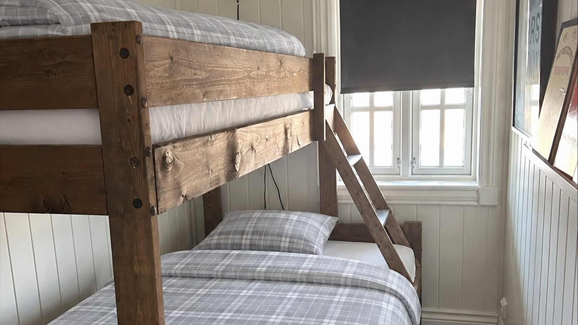Soveområde med køyeseng i tre, grå rutete sengetøy og vinduer som slipper inn naturlig lys.