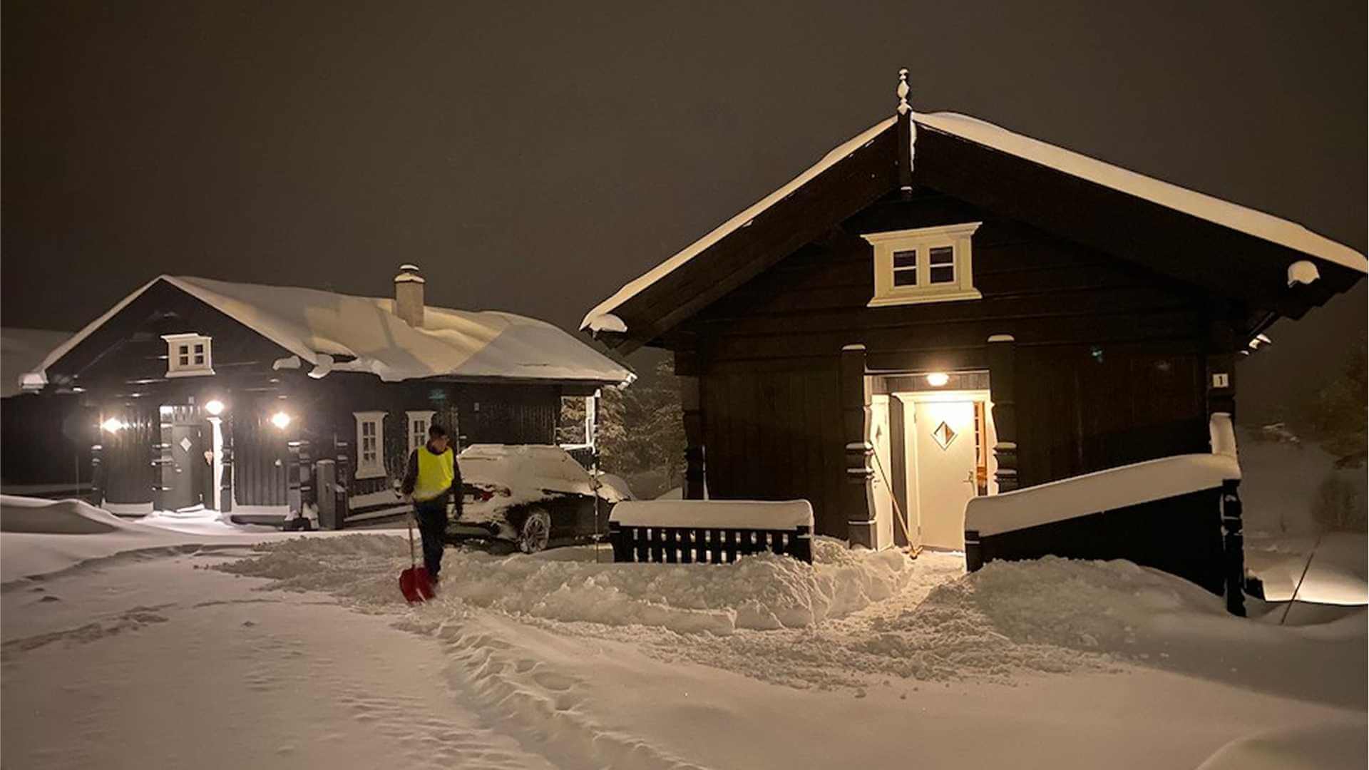 En mann som måker snø foran to sorte hytter under et stjerneklar natt med snødekt landskap.