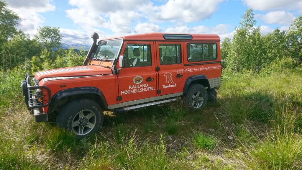 Rød offroad-kjøretøy merket Rauland Høgfjellshotell i norsk natur med grønt landskap og blå himmel.