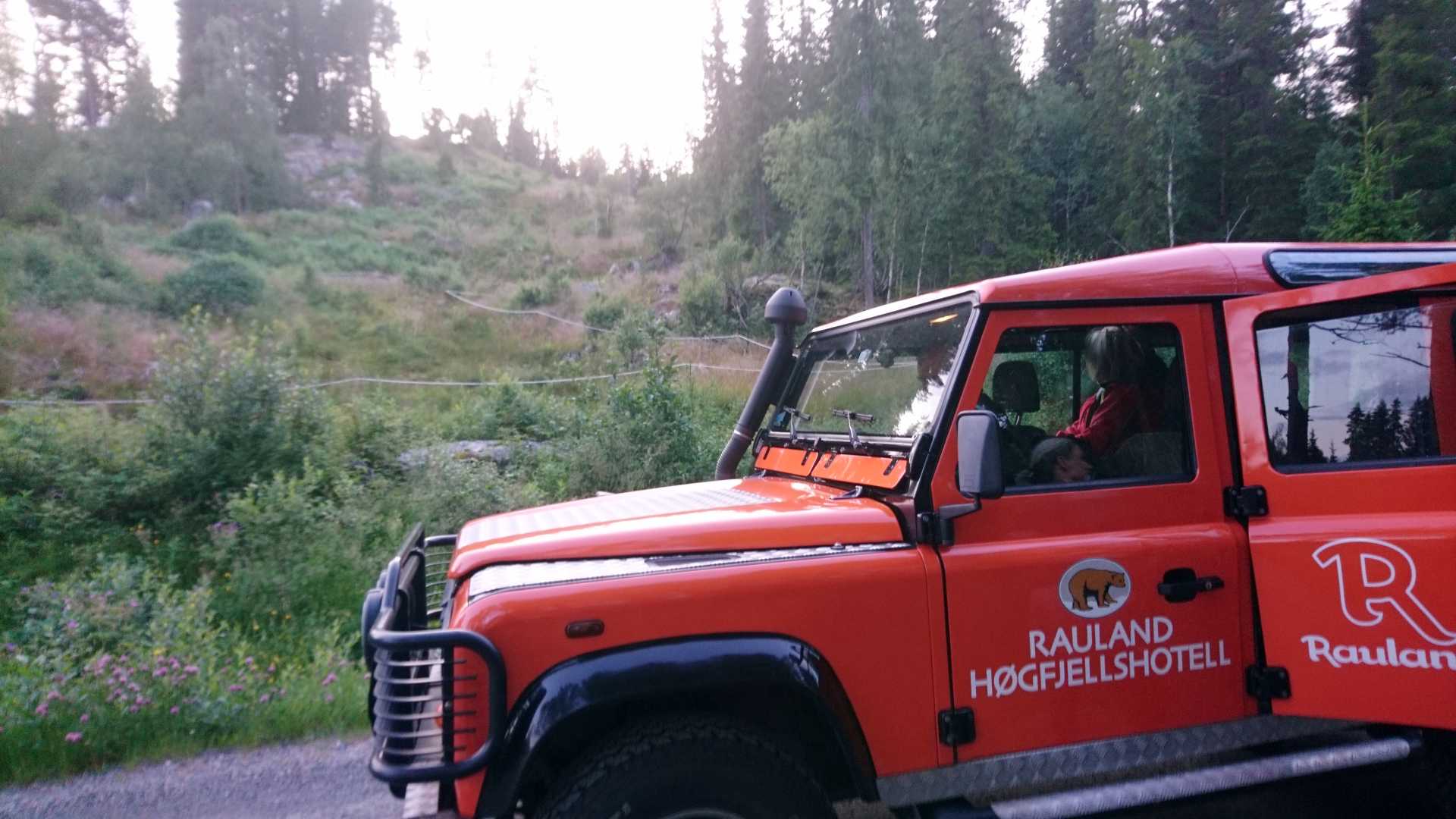 Rød bil fra Rauland Høgfjellshotell kjører gjennom skogkledd område i Norge.
