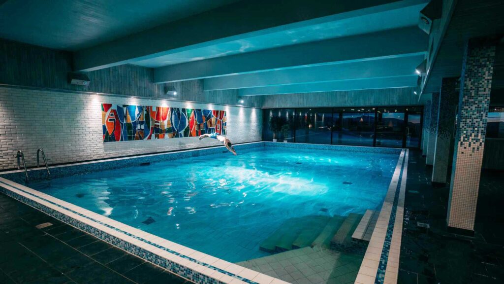 Person som stuper i et opplyst innendørs svømmebasseng med fargerik veggdekorasjon.