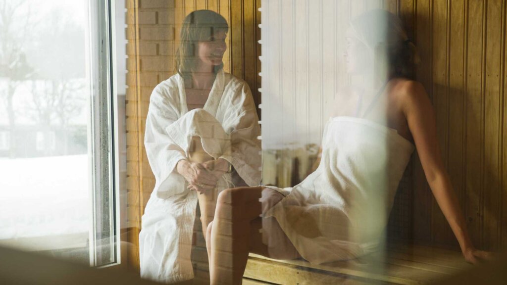 To personer slapper av i en sauna, kledd i håndklær, med utsikt til vinterlandskap gjennom vinduet.