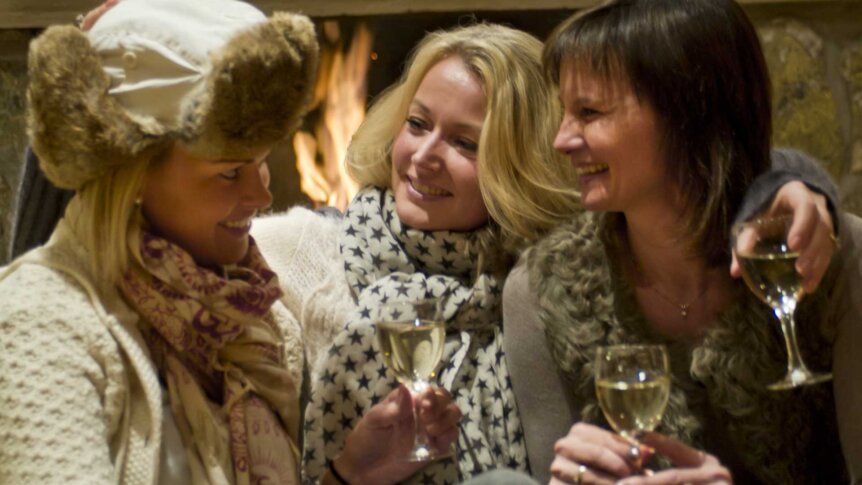 Tre kvinner nyter vin ved peisen i vinterklær og skjerf, med smil og vennskap.