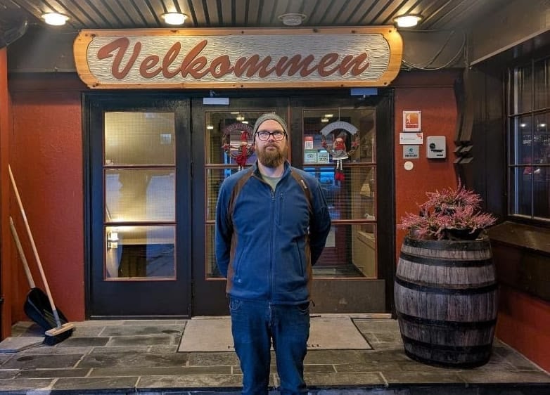 Mann foran inngang med Velkommen-skilt, kledd i blå jakke, lue, ved siden av tønne og kost.