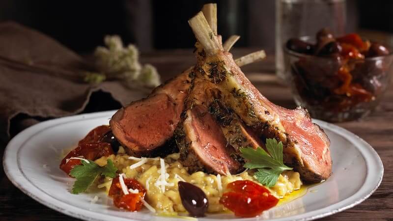 Grillet lammekoteletter med risotto, urter og oliven, servert på hvit tallerken i rustikk setting.