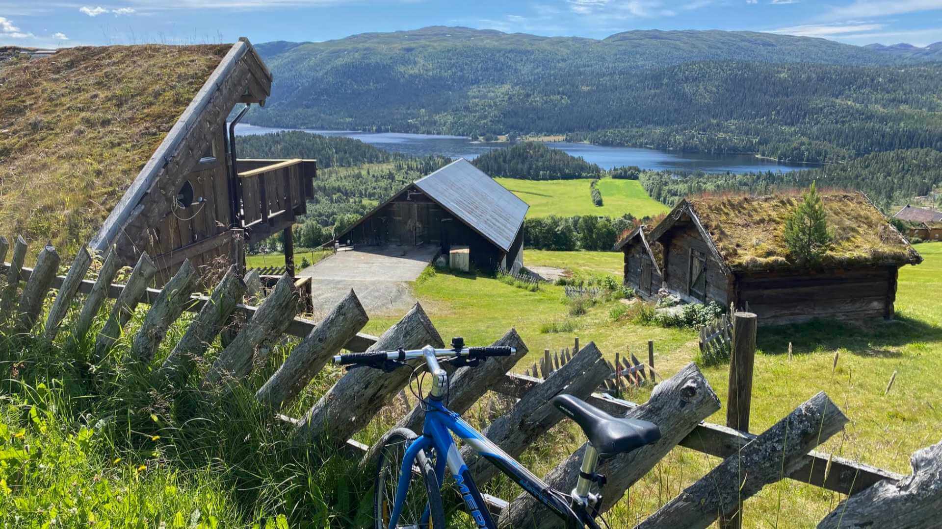 Bilde av en blå sykkel foran tradisjonelle hytter med grønt tak, omgitt av vakker norsk natur og fjellutsikt.