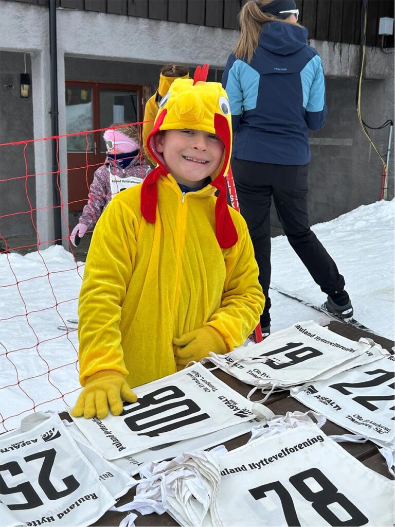 Barn i kyllingkostyme smiler mens de holder på med skiarrangement, med nummerlapper på bordet.