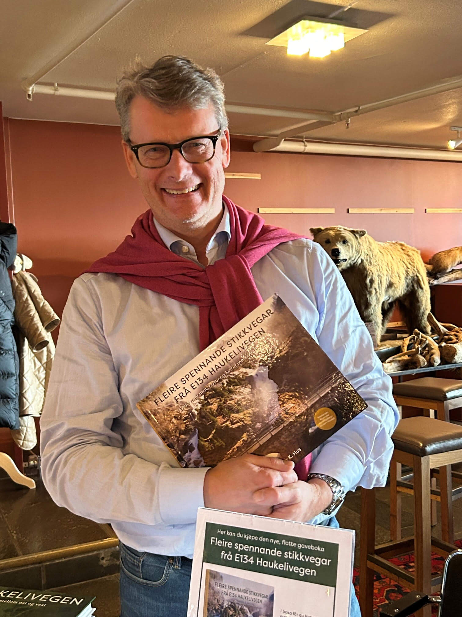En smilende mann holder en bok om Haukelivegen, med en bjørn i bakgrunnen. Boken viser spennende stier i området.