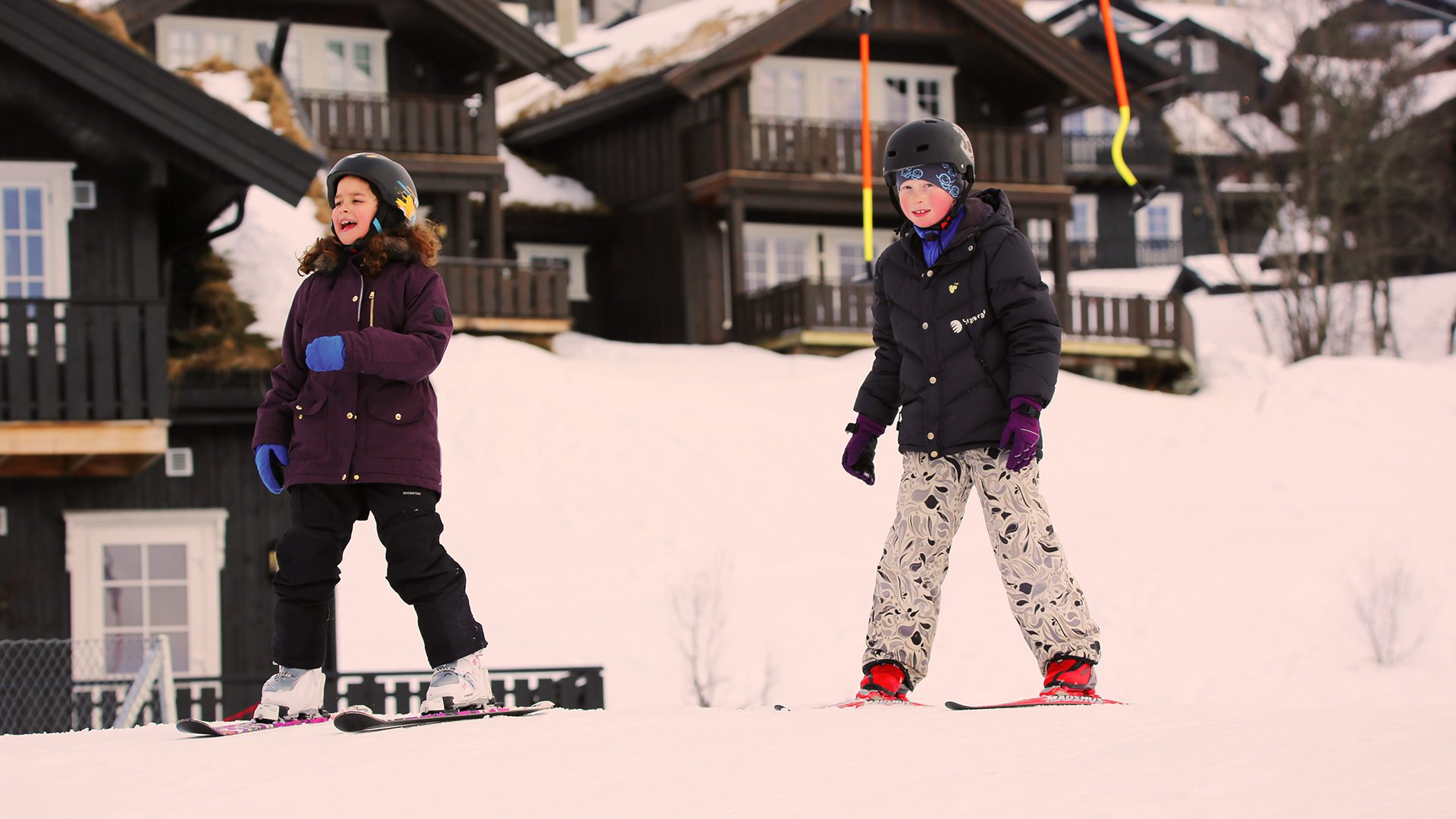 To barn p skitur i sndekt skisted, ifrt vinterklr og hjelmer, med hytter i bakgrunnen.