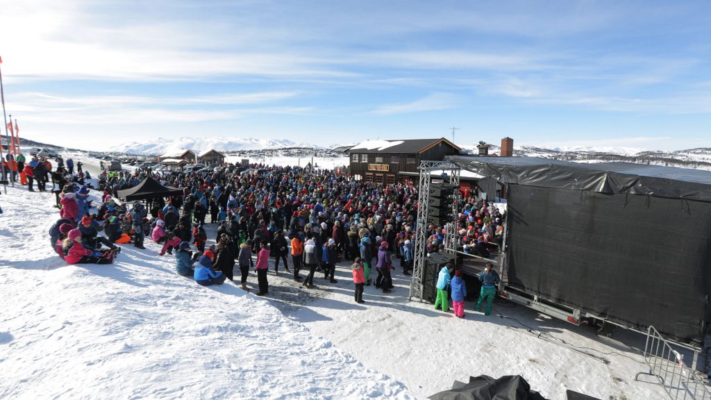 Stor folkemengde nyter utendørs konsert i et snødekket fjellandskap, solfylt dag med blå himmel.