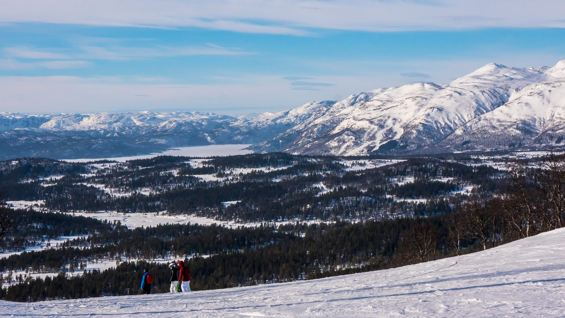 Tre personer i fargerike klær går på snødekte fjell med vakker utsikt over dalen og fjellene i bakgrunnen.